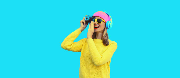 时尚的现代年轻女摄影师戴着彩色的粉红色帽子，黄色的毛衣，蓝色的工作室背景，用胶片相机拍照，戴着耳机听音乐图片下载