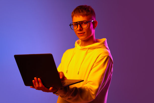 年轻人，戴眼镜的学生，拿着笔记本电脑站在霓虹灯下渐变的紫色背景下图片下载