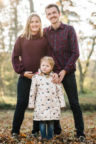 年轻的家庭在大自然中共度时光。怀孕的妈妈和爸爸站在森林的金色树叶背景上拥抱女儿。夕阳下，幸福的一家人在秋天的公园散步。特写镜头。秋天的照片。图片下载