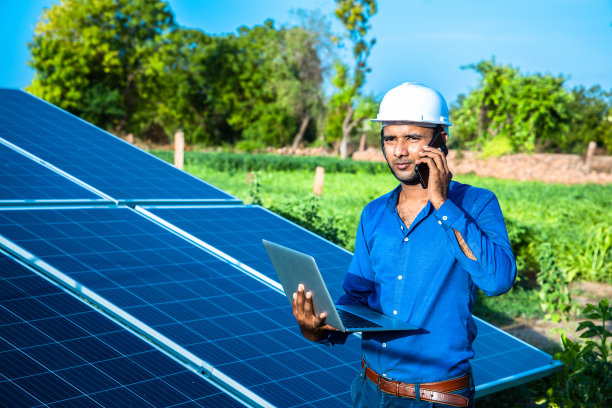 年轻的男工程师拿着笔记本电脑站在太阳能电池板旁边讲电话，农业农场有清澈的蓝天背景，可再生能源，清洁能源。图片下载
