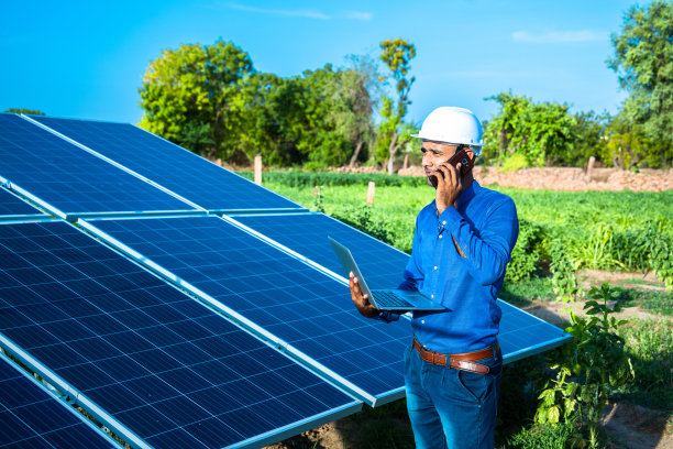 年轻的男工程师拿着笔记本电脑站在太阳能电池板旁边讲电话，农业农场有清澈的蓝天背景，可再生能源，清洁能源。图片下载