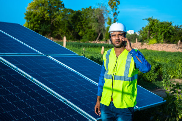 穿着绿色背心和头盔的年轻男技术工人在电话中谈论太阳能电池板的维护和安装，站在田间，农业技术图片下载