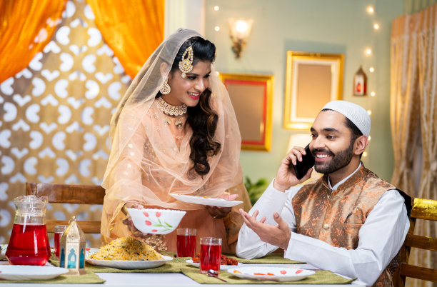 快乐的印度丈夫打电话给朋友或亲戚，而妻子在节日庆祝活动中安排斋月晚餐或盛宴——技术、团聚和斋月问候的概念图片下载