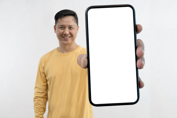 一个满意的、自信的、微笑着的男人站在工作室的复印空间里，向相机展示着手机的空白屏幕模型。图片下载