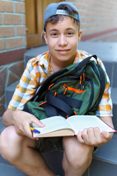 学校的学生，一个十几岁的男孩正坐在楼旁的台阶上，读着一本书，一个明媚的夏日，回到学校和教育理念图片下载