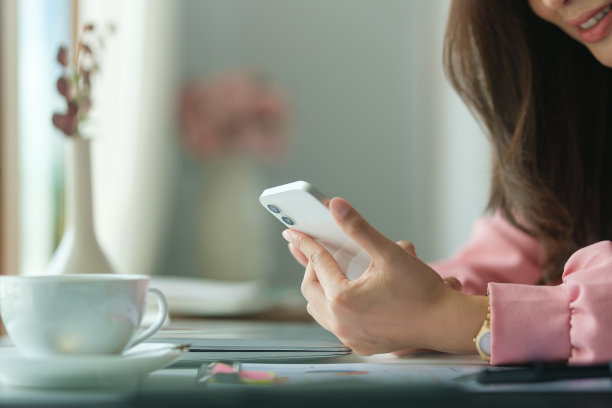剪短的照片，微笑的年轻女商人坐在她的办公桌前，使用手机。图片下载