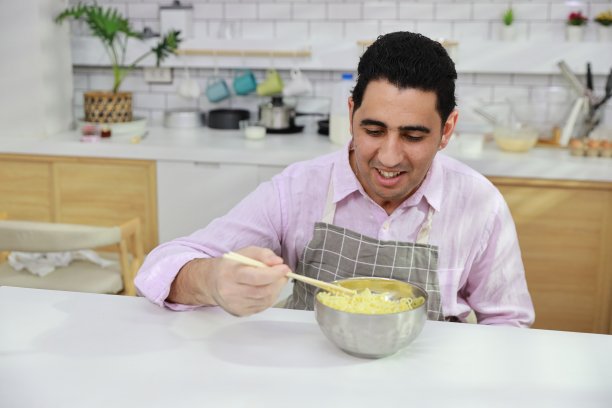 特写年轻快乐的西班牙卷发男人坐在桌子上吃美味的方便面午餐在厨房用筷子和砖墙。不健康和廉价食品的生活方式概念图片下载