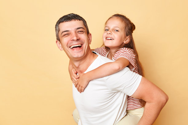 开朗的孩子背着深褐色头发的父亲，孤立在米色背景下，一家人在一起开心地笑着，穿着休闲服，心情很好。图片下载