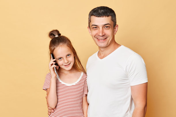 开朗快乐的男人和小女孩站在一起，孤立在米色的背景下，可爱的女儿在用智能手机聊天，父亲带着快乐的脸看着相机。图片下载