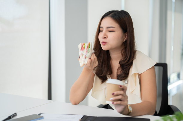 一位美丽而饥饿的亚洲女商人坐在办公桌前吃着三明治，喝着咖啡图片下载