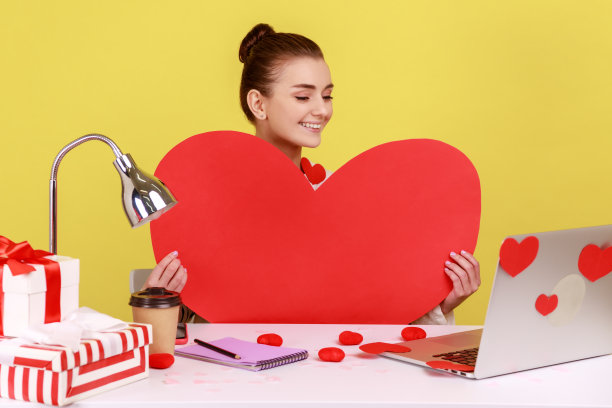 拿着红纸心的女人，坐在工作场所，用粘心的笔记本电脑，进行视频通话。图片下载