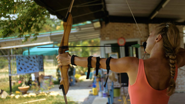 在射击场拿弓的女运动员。射击场的运动员瞄准箭以射中目标。户外射箭训练。射击场中射箭的练习和训练。运动员保持木弓。图片下载
