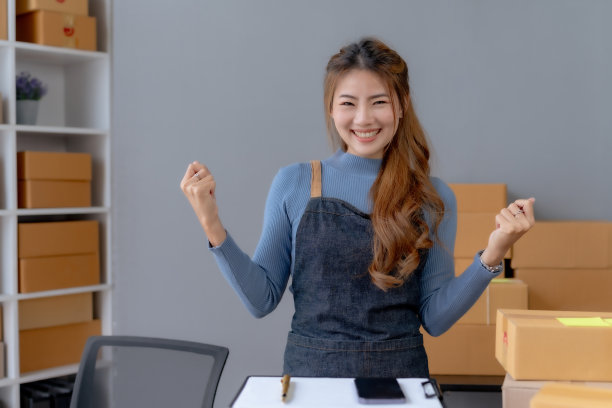 成功的亚洲女企业家在家里对着笔记本电脑惊喜地举手微笑。小企业主亚洲女性谁是高兴的成功看着镜头。图片下载