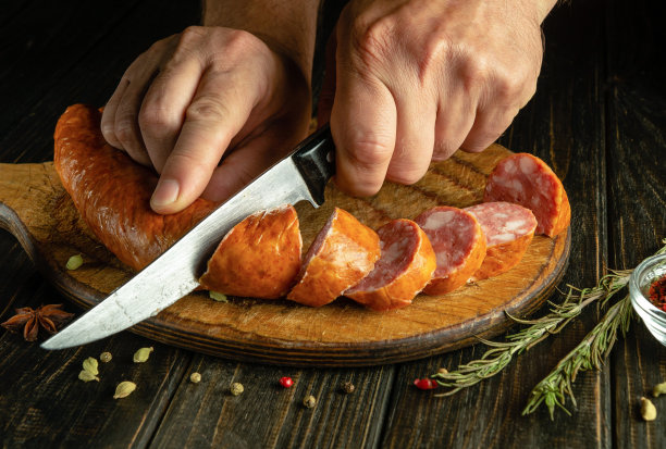 在家做香肠三明治的概念。一个男人拿着刀在厨房板上切肉肠的手图片下载