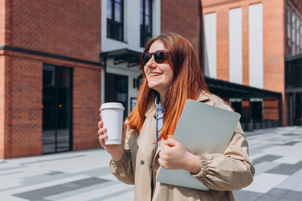 白种红发女商人，穿着时尚的办公室服装，拿着笔记本电脑和生态茶在外面摆姿势。模型，女人用笔记本电脑和拿着生物白色的咖啡杯图片下载