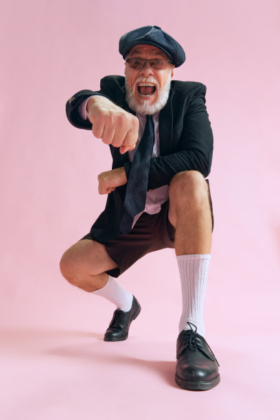 老年大胡子男子的俯视图照片，穿着衬衫黑色短裤漂亮的太阳镜和帽子，坐在粉红色的背景上孤立的拳头图片下载