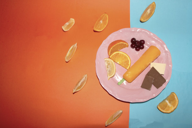 粉红色盘子里放着樱桃、桔子片、巧克力和桔子冰淇淋，背景是蓝橙，创意夏季概念，平面图片下载