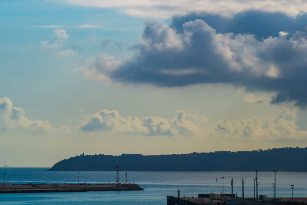 令人惊叹的美丽的海岸景色与港口防波堤。大气的图片，海洋，山脉，各种颜色的云在阳光下的阴影，背景是一个几乎不明显的帆船图片下载