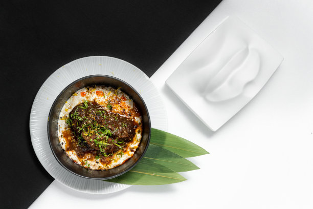 红烧小牛肉配上大葱和土豆泥。食物放在一个深色的陶瓷碗里，放在一个装着竹叶的浅盘子里。这些盘子立在一张黑白的底纸上。图片下载