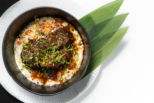 红烧小牛肉配上大葱和土豆泥。食物放在一个深色的陶瓷碗里，放在一个装着竹叶的浅盘子里。这些盘子立在一张黑白的底纸上。图片下载