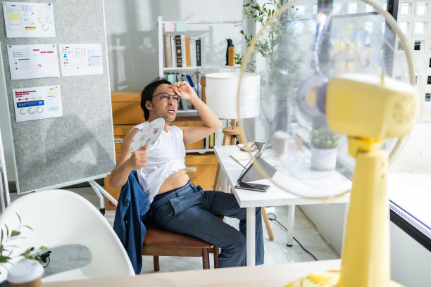 商务人士一边拿着文件和笔记本电脑坐在办公室里，一边用电风扇给自己吹气。图片下载