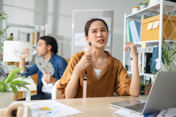 商务人士坐在办公室里拿着文件和笔记本电脑，一边用电风扇吹自己。图片下载