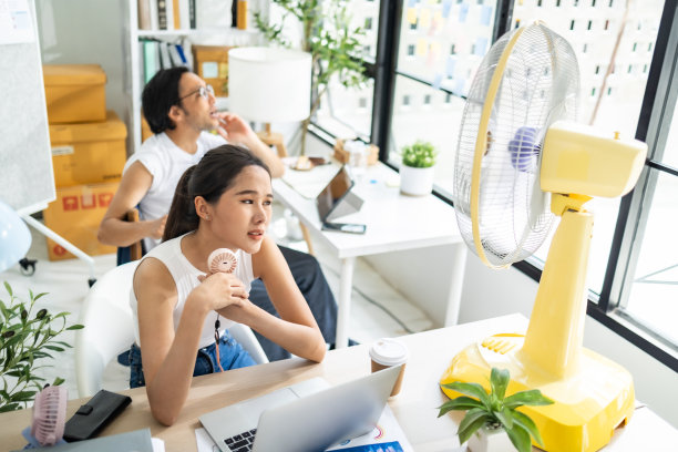 亚洲商务人士坐在办公室里拿着文件和笔记本电脑，一边用电风扇吹自己。图片下载