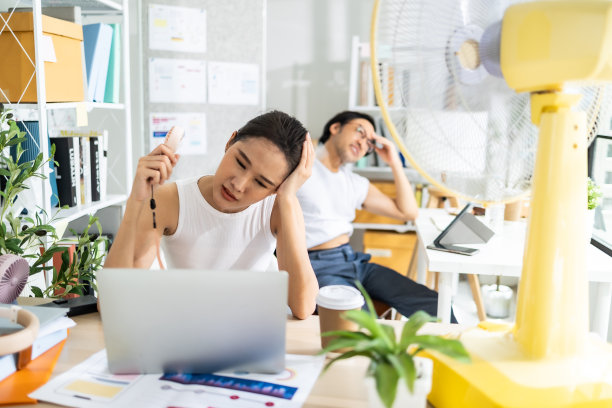 亚洲商务人士坐在办公室里拿着文件和笔记本电脑，一边用电风扇吹自己。图片下载