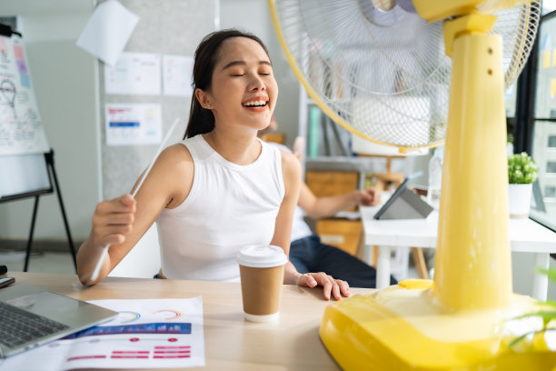 亚洲女商人喜欢一边吹电扇，一边坐在办公室拿着文件和笔记本电脑。图片下载