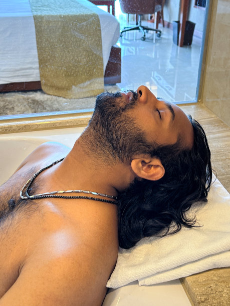 特写照片:印度男子躺在白色浴缸里，闭着眼睛，在浴缸里放松，头和肩膀靠在浴盆边的毛巾上，毛茸茸的胸部，面部毛发，头像，侧面视图图片下载