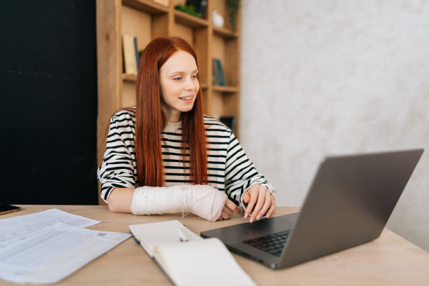 积极的女性自由职业者的肖像，右手骨折，用石膏绷带包裹着，坐在桌子旁，在笔记本电脑上工作，微笑着看着屏幕图片下载