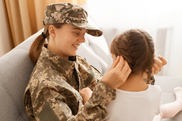 侧面照片中，身着迷彩服、头戴迷彩帽的女子与女儿合影，母亲从战场或军队归来，带着爱和微笑看着孩子。图片下载