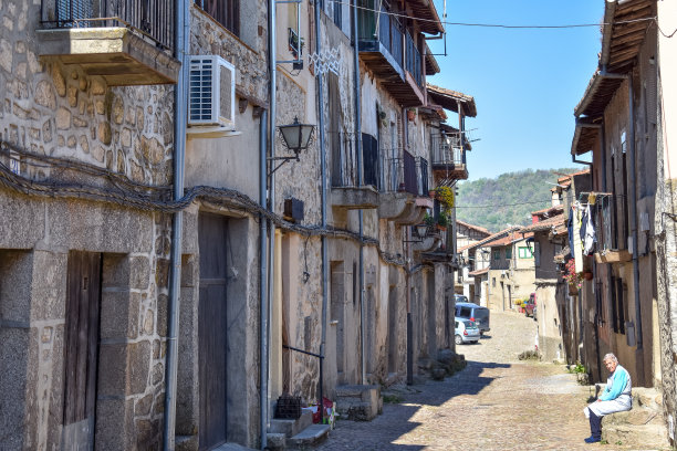 一位老妇人独自坐在长椅上，在空旷的街道上享受着阳光明媚的一天。它反映了西班牙农村地区由于缺乏就业而人口减少。图片下载