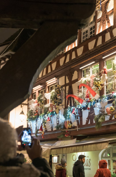 在法国历史悠久的斯特拉斯堡，一位老人在传统的圣诞市场上用手机拍下装饰精美的半木房子的照片图片下载