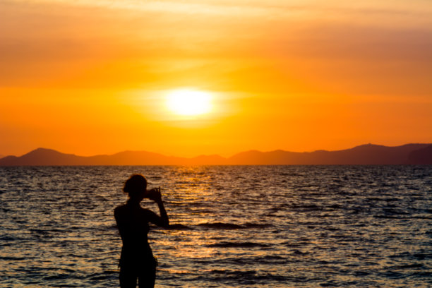 剪影女子使用相机手机拍照在海滩上日落在海上背景。模糊的照片图片下载