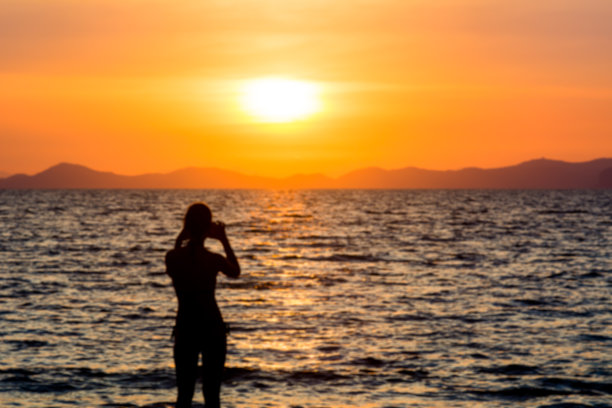 剪影女子使用相机手机拍照在海滩上日落在海上背景。模糊的照片图片下载
