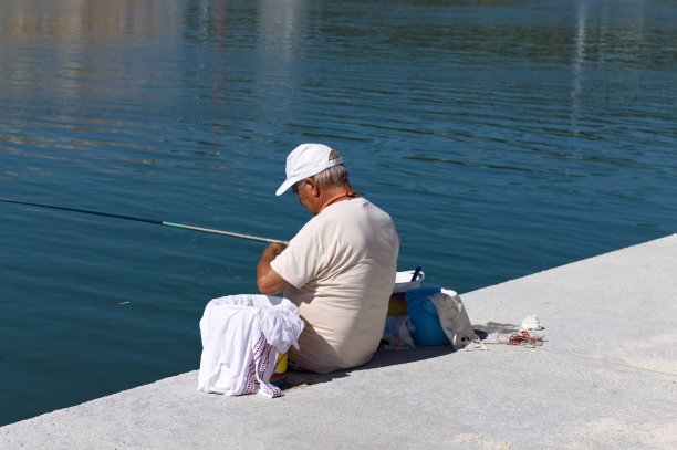 一位戴着白帽的老渔夫正在用他的钓竿在港口的码头上钓鱼(意大利，佩萨罗)图片下载