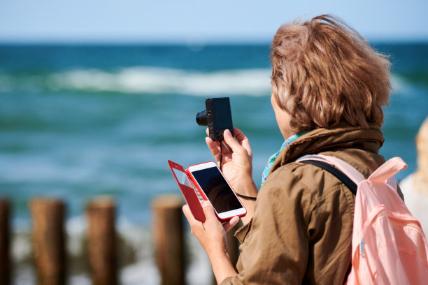 棕色头发的女人在海上拍照，用数码相机拍摄，拿着她的智能手机图片下载