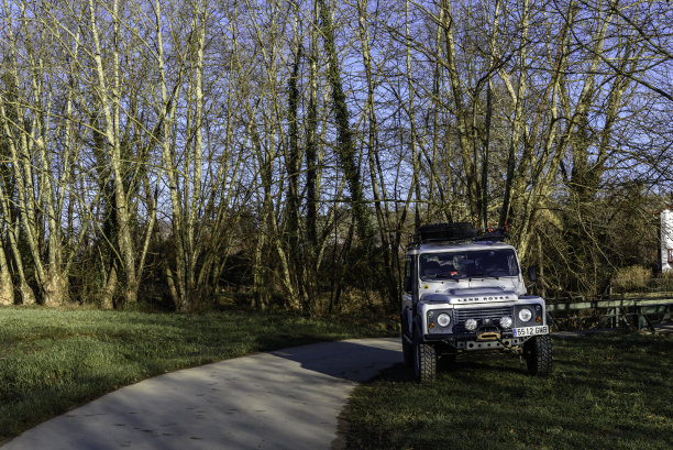 前面的照片是一个白色的路虎防守者从一边看向右边的照片，在路边的草地上，后面是一排光秃秃的树木，在秋天图片下载