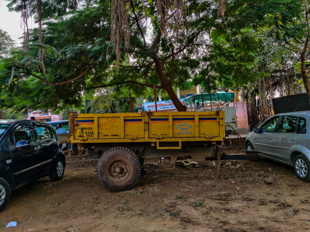 库存照片黄色油漆的牵引车挂车停在停车场的树荫下，灰色和深蓝色的汽车停在挂车的旁边。图片下载