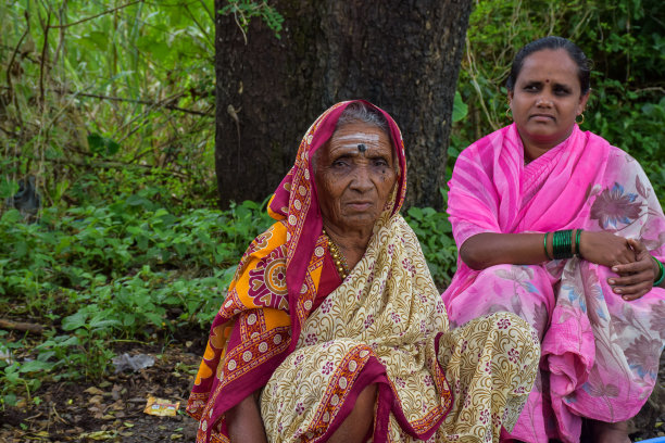 照片中，两名40至70岁的印度妇女穿着纱丽坐在路边，清晨在公交车站等公交车或公共交通工具。图片下载