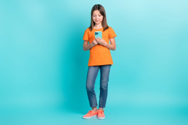 可爱的漂亮女孩女儿的全身照片沉迷于用户穿着时尚的橙色衣服拿着iphone孤立在青色背景图片下载