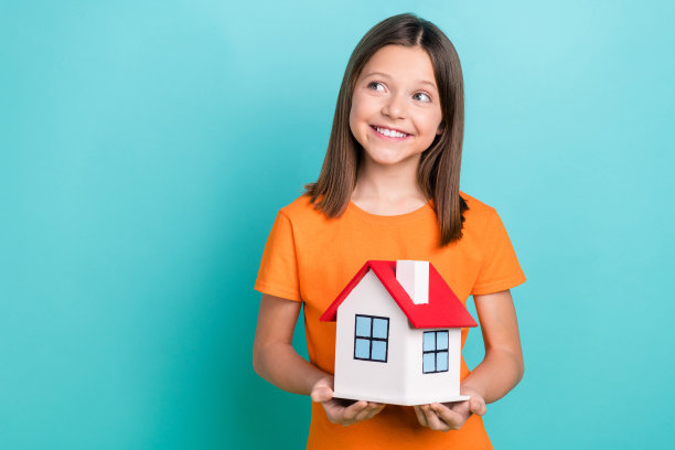 照片中漂亮满意的小女孩穿着橙色时髦的t恤，双手捧着小房子，在蓝绿色的背景上看起来空无一物图片下载