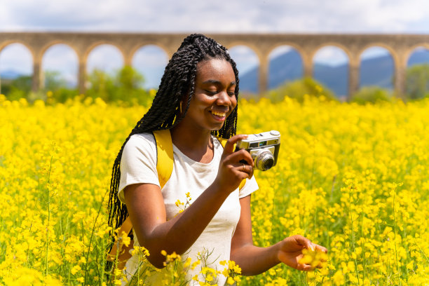 一个扎着辫子的黑人女孩，一个旅行者，在一片黄花中，用老式相机拍着花的照片图片下载