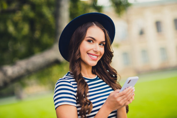 漂亮可爱的年轻女士戴条纹裙头饰微笑行走打字现代设备户外城市城市公园的照片图片下载