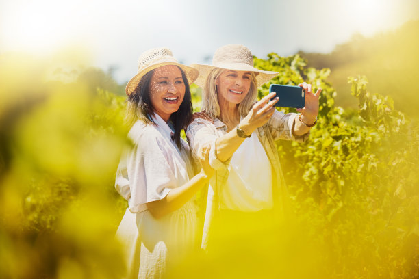 两个快乐的朋友在葡萄园里用手机自拍。白天，白人和混血女性微笑着站在一起，用手机拍社交媒体上的照片图片下载
