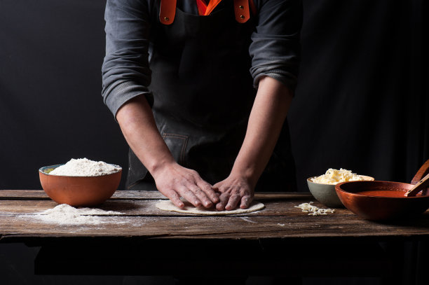 照片上，一名男子在一个老式的木制柜台上用手揉着披萨面团，上面放着一碗面粉、番茄酱和奶酪图片下载