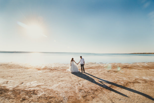 新婚夫妇在海边散步。阳光明媚的夏日照片。新娘披散着头发，穿着露肩裙。海洋浪漫的仪式。海边的爱情故事。沙子，水和地平线。蜜月。图片下载