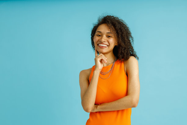 穿着橙色连衣裙的漂亮女人微笑着抚摸着脸颊，看着蓝色背景的相机图片下载