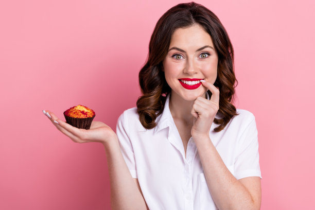 照片中可爱狡猾的女人穿着白衬衫，手指，嘴唇，嘴巴，手，手臂小纸杯蛋糕孤立的粉红色背景图片下载